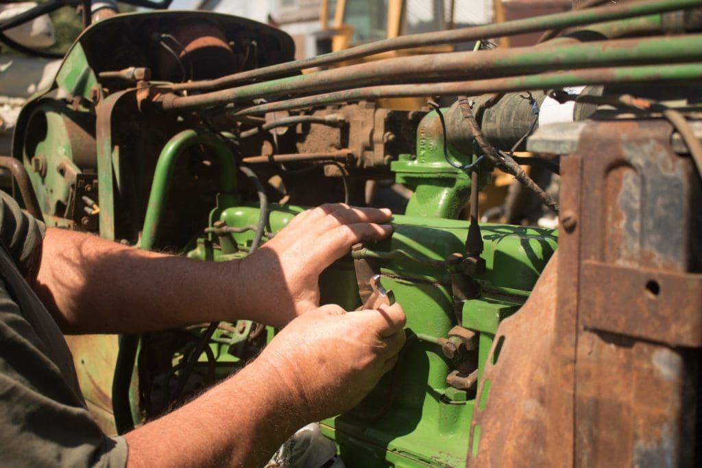 A repairman working on an old green tractor attemping to repair mechanical wear and tear.